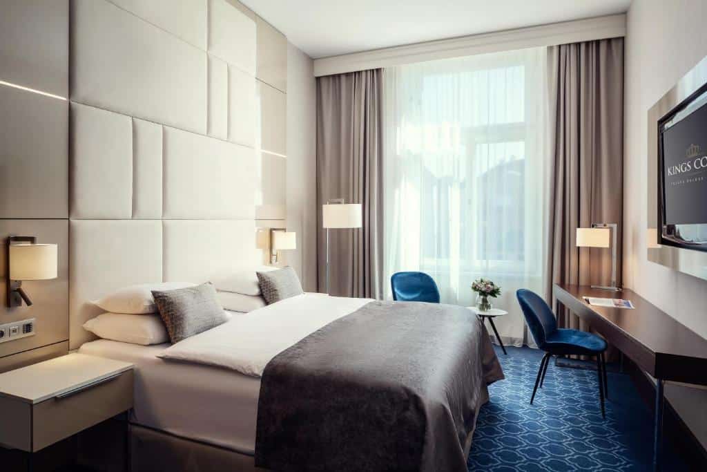 Quarto de hotel moderno do Hotel Kings Court, com cama de casal, duas mesas de cabeceira, cadeiras azuis, uma secretária e uma TV na parede.