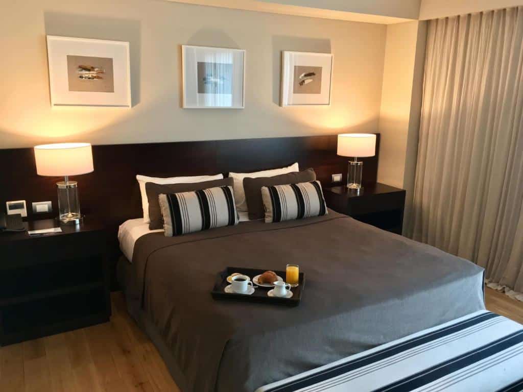 quarto do Hotel Metropolitano Supara com cama de casal ao centro e um pequeno-almoço servido em cima, mesas de madeira e candeeiros dos dois lados e três quadros na parede, em tons acastanhados
