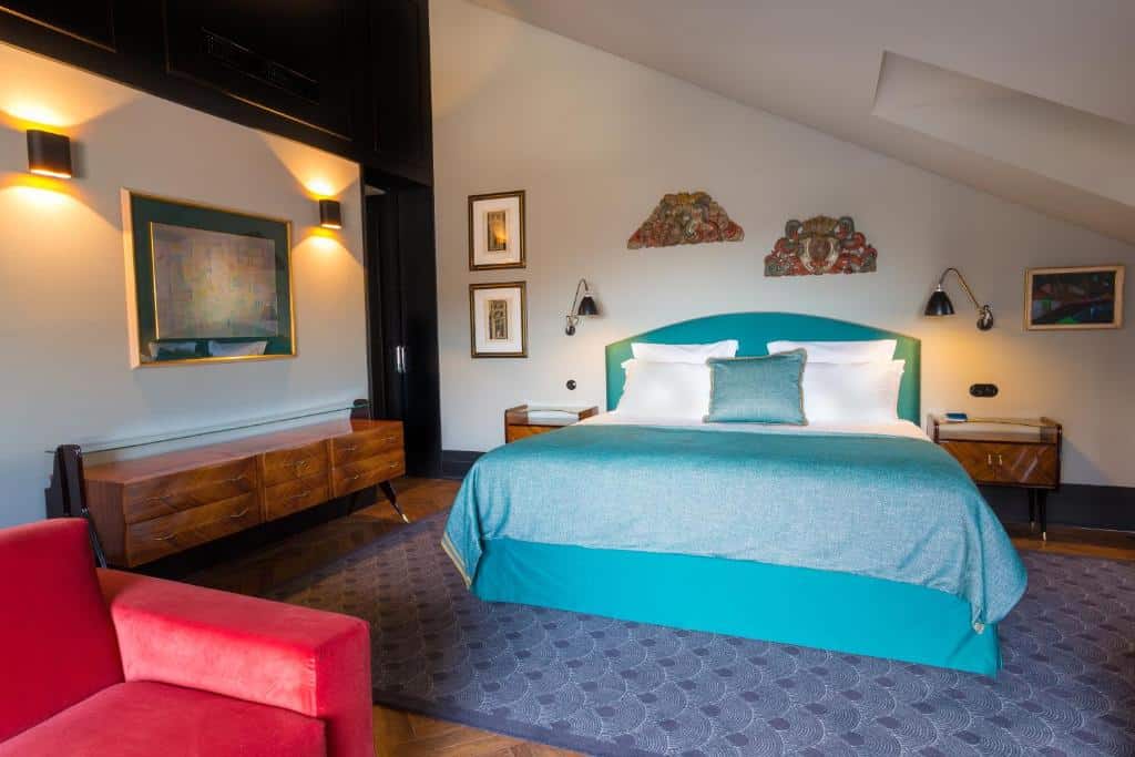 Quarto do Hotel Valverde Lisboa, com cama de casal e almofadas, à esquerda uma cómoda de época em madeira e um quadro, uma poltrona vermelha e duas mesinhas de madeira de cada lado da cama