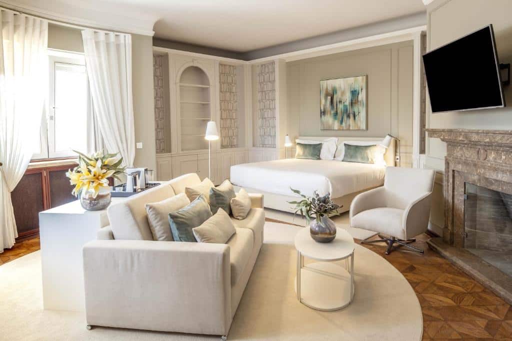 Quarto espaçoso do Luzeiros Suites com uma cama de casal, um pequeno sofá de dois lugares com várias almofadas em frente a uma televisão e, do lado esquerdo, uma janela e um móvel com utensílios para preparar café.