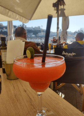Uma bebida vermelha gelada do Botequim Nostalgic, decorada com uma rodela de limão e uma palhinha preta, sobre uma mesa de madeira num café ao ar livre. Representa o que fazer à noite no Porto.