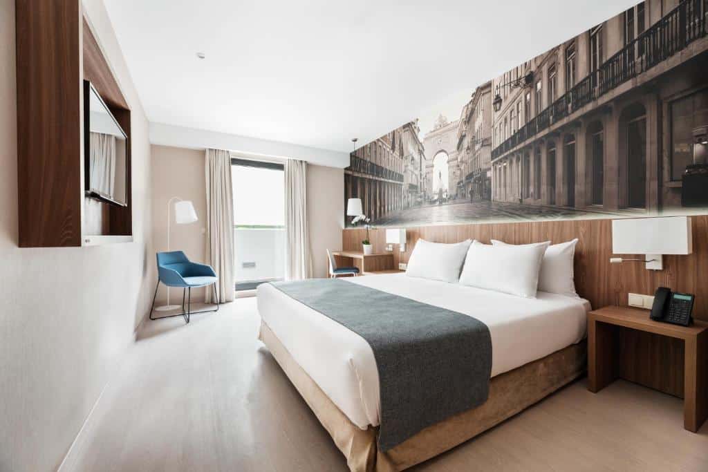Quarto do Exe Saldanha com pequena varanda com cortinas, cama de casal com almofadas, secretária do lado esquerdo e uma televisão na parede em frente à cama