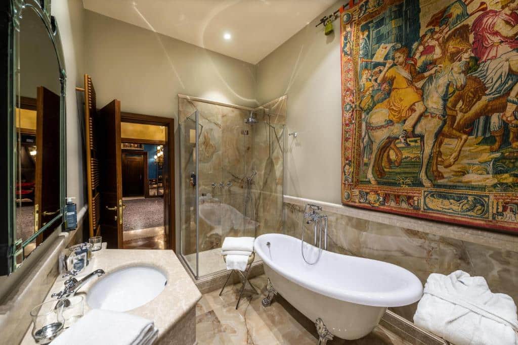 Casa de banho luxuosa com banheira de porcelana branca, chuveiro de vidro e paredes decoradas com uma enorme tapeçaria em tons quentes que retrata uma cena de cavalaria antiga
