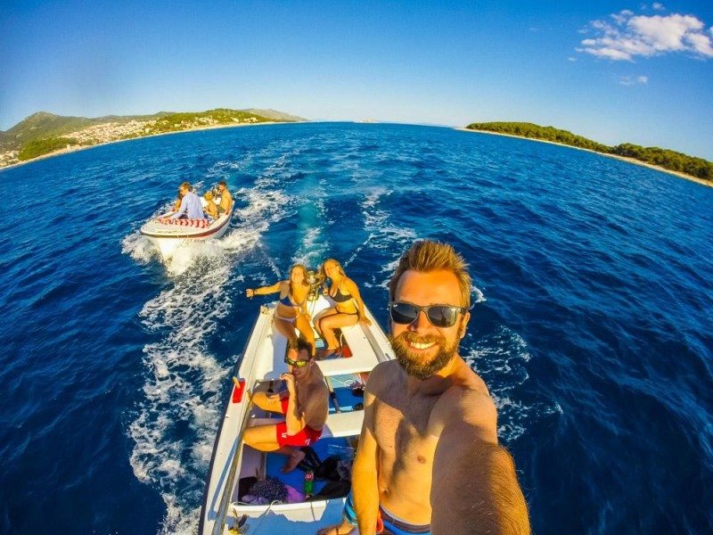 Pessoas a sorrir num barco no mar, rumo às Ilhas Pakleni, na Croácia, com uma a tirar uma selfie. Ao fundo há outro barco com pessoas.