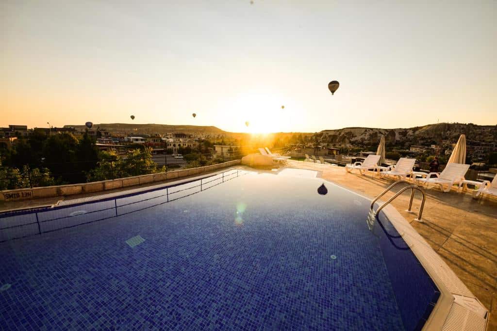 Piscina exterior ao nascer do sol no Cappadocia Caves Hotel, com espreguiçadeiras, chapéus de sol e balões de ar quente visíveis no céu sobre as colinas.