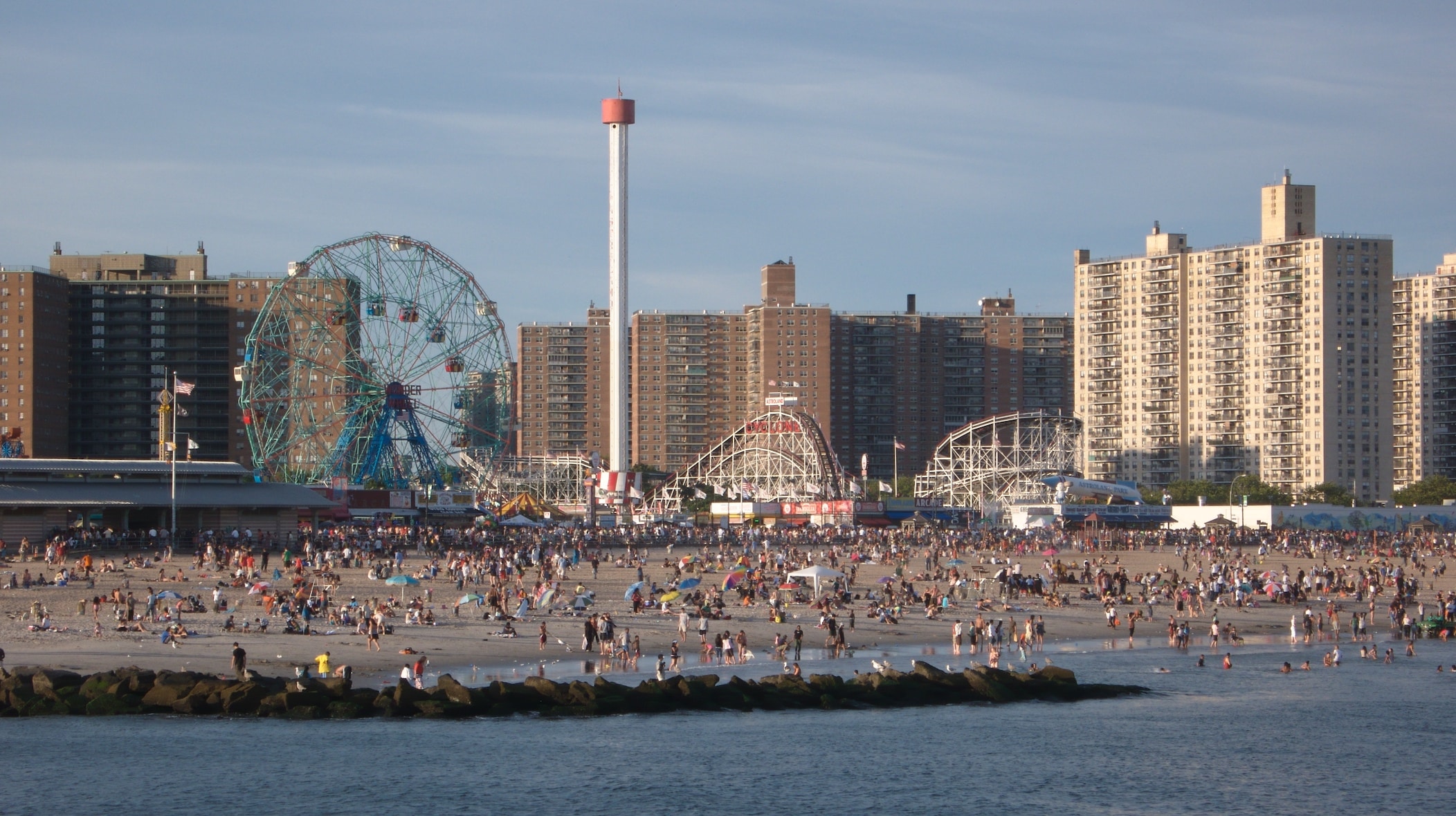 Praia cheia de gente em Coney Island, com uma roda-gigante e montanhas-russas de um parque de diversões ao fundo.