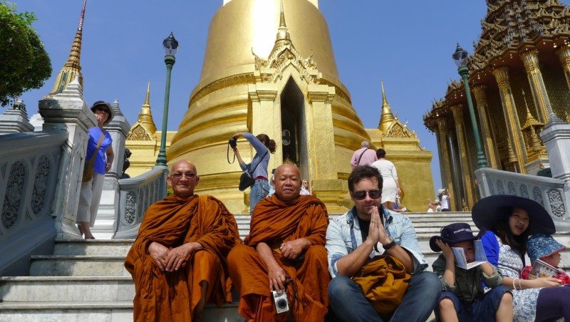 Monges sentados nos degraus do Grand Palace em frente a um pagode dourado, num dia claro e soalheiro.