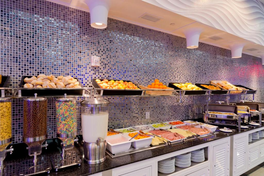 Zona de pequeno-almoço do Riu Plaza com uma variedade de alimentos num buffet: pão, cereais, fruta e outros itens, bem organizados. A parede ao fundo é decorada com azulejos brilhantes.