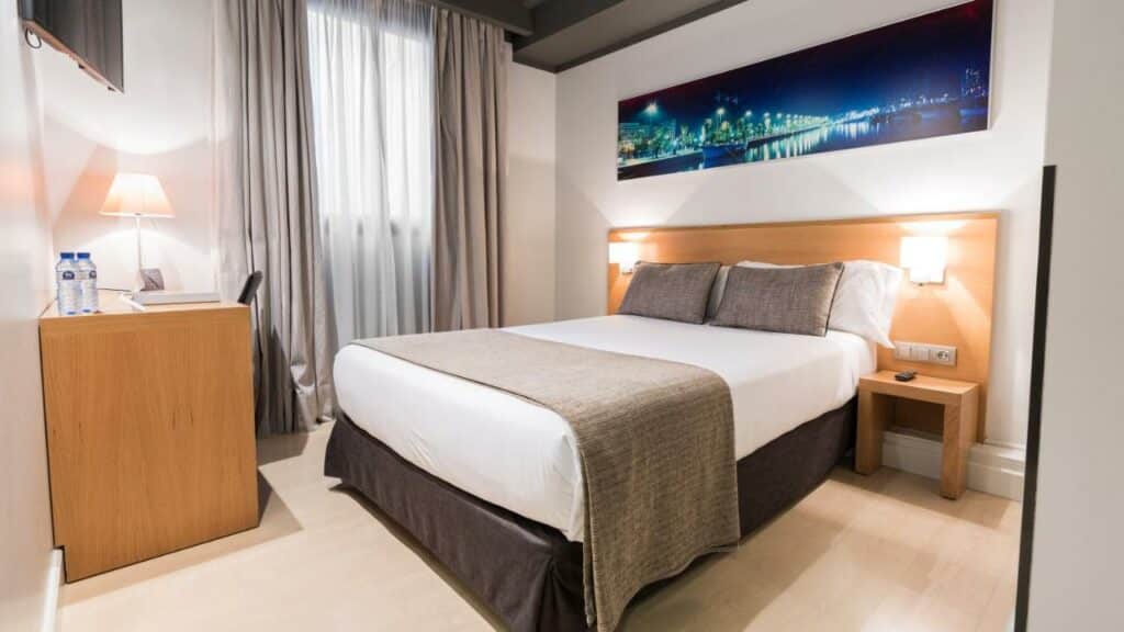 Quarto do Arc la Rambla, com cama de casal, candeeiros fixos na cabeceira de madeira, televisão virada para a cama e secretária junto a uma janela com cortinas bege.
