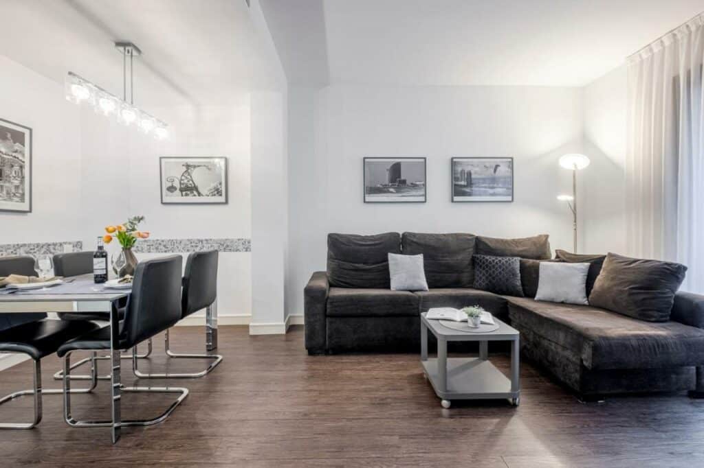 Interior de um apartamento do Catedral Bas Apartments. À direita, uma sala de estar com sofá e mesa de centro; à esquerda, uma sala de jantar com mesa posta e quatro cadeiras.