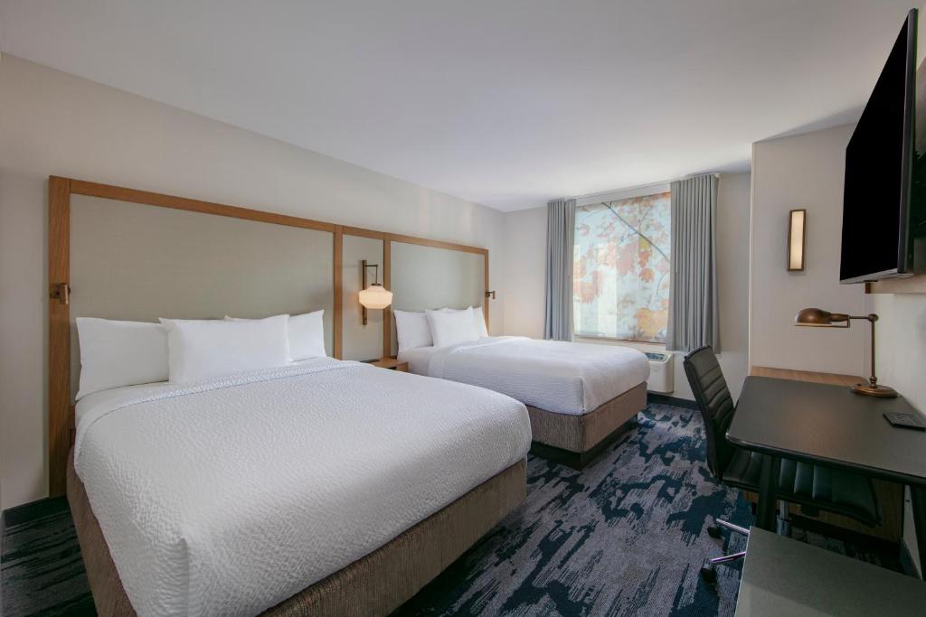 No quarto do Fairfield Inn by Marriott JFK Airport há duas camas de casal com cabeceiras estofadas ao centro. À direita, uma secretária com cadeira preta e uma TV na parede. À esquerda, uma janela com cortinados cinzentos e, a um canto, uma poltrona verde.