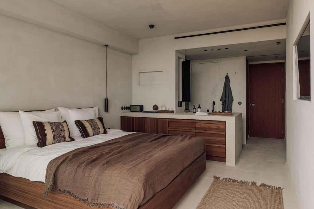 Quarto minimalista do OKU Ibiza com colcha castanha, mobiliário de madeira e paredes brancas.