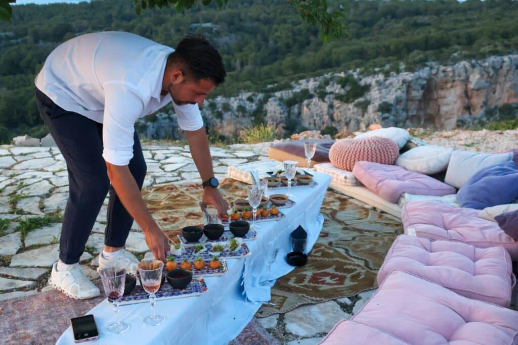 Um homem prepara comida e bebidas numa mesa baixa num pátio de pedra ao ar livre em Hvar, com vista panorâmica e assentos confortáveis à volta.