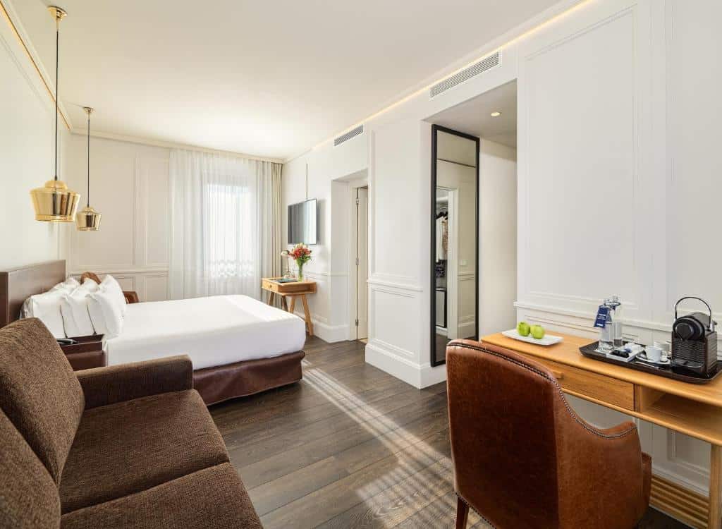 Quarto do Boutique Hotel H10 Montcada. A cama ao fundo tem candeeiros nas mesas de cabeceira, televisão em frente, uma janela ao lado e um sofá castanho com mesa e máquina de café.