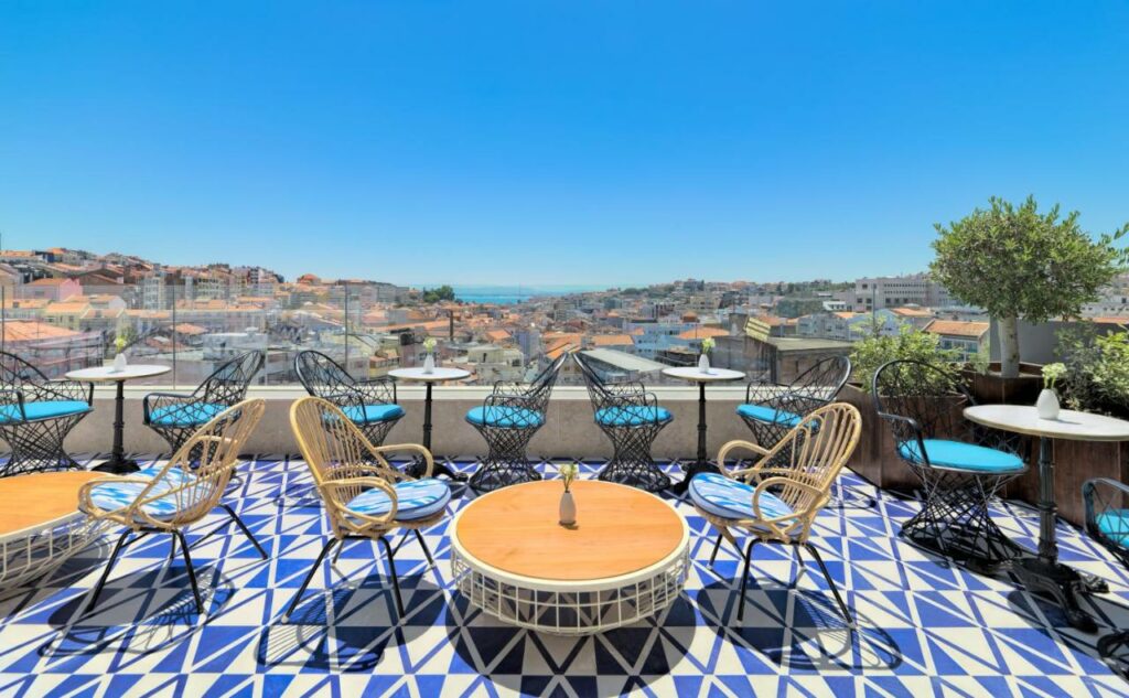 Terraço ao ar livre com pavimento padronizado em azul e branco, cadeiras de vime, mesas pequenas e vista sobre a cidade, no H10 Duque de Loulé.