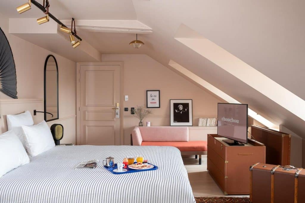 Cama do Chouchou Hotel com colcha às riscas, tabuleiro de pequeno-almoço, sofá cor-de-rosa e teto inclinado num quarto moderno.