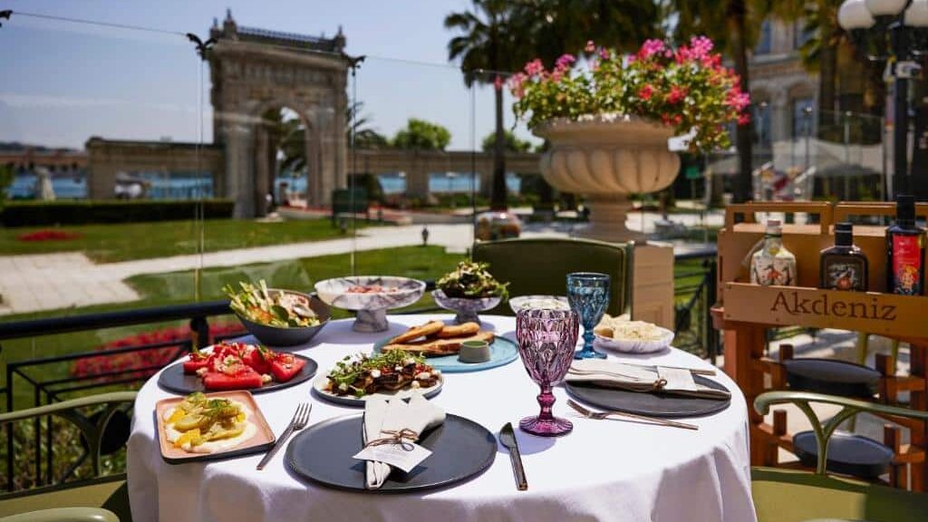 Uma mesa redonda do Çırağan Palace Kempinski Istanbul para duas pessoas, com vários pratos e bebidas, num terraço com vista para um arco de pedra histórico e um jardim.