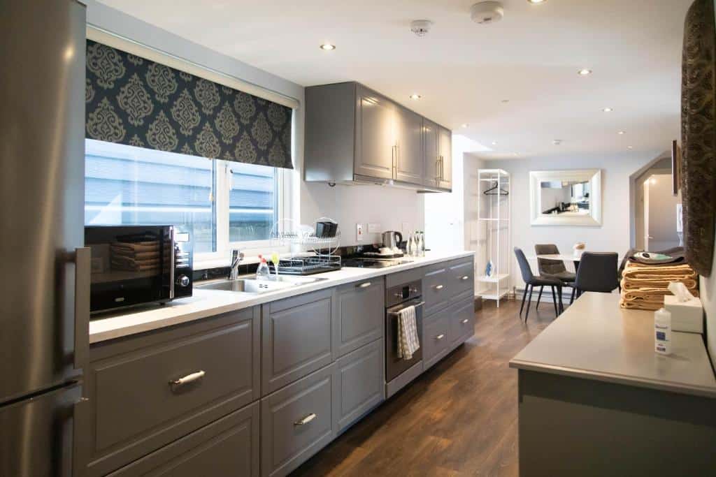 Cozinha moderna do Molloys Apartments com armários cinzentos, bancada clara, eletrodomésticos e utensílios, com vista para a sala de estar com cadeiras escuras.