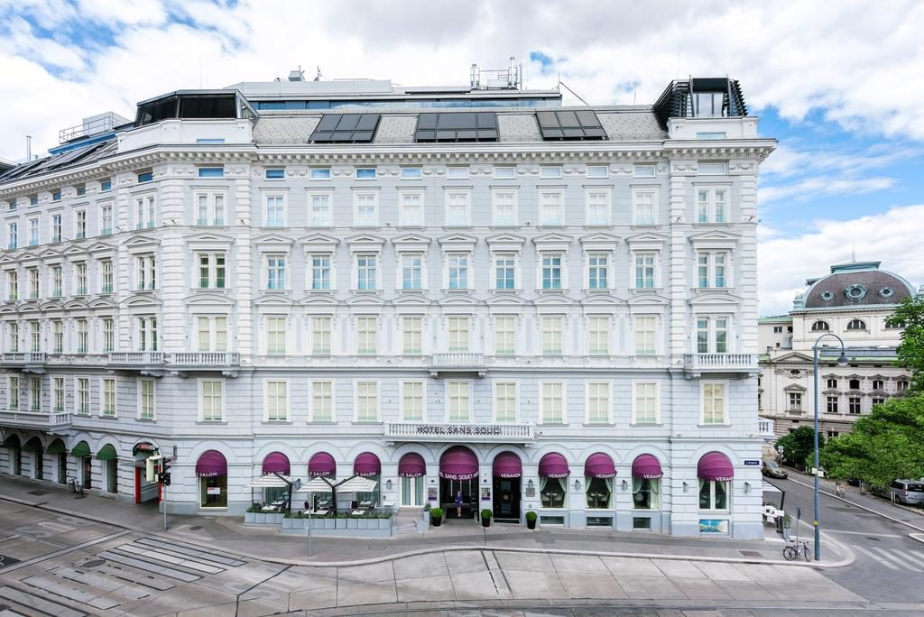 Grande edifício do Hotel Sans Souci Wien, branco, com toldos roxos, numa rua da cidade sob um céu parcialmente nublado.