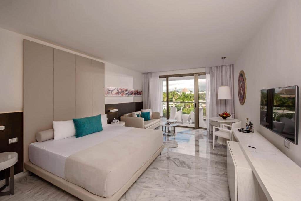 Quarto do Platinum Yucatan Princess Adults Only com cama de casal ao centro, sofá bege ao fundo e vista para área verde pela varanda. Em frente à cama há uma TV.