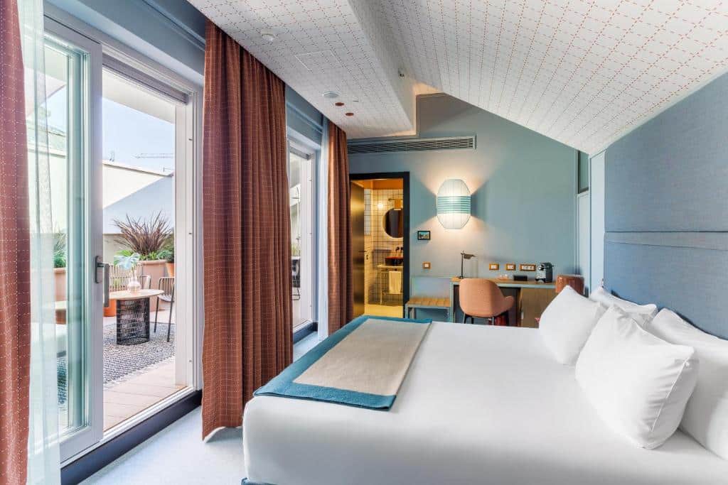 Quarto do Room Mate Giulia com varanda ampla, mesinhas e cadeiras, vasos de flores, cama de casal e secretária
