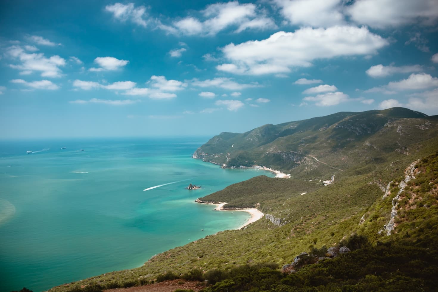 Vista aérea de uma paisagem costeira com mar azul-turquesa, praias de areia e colinas verdes.