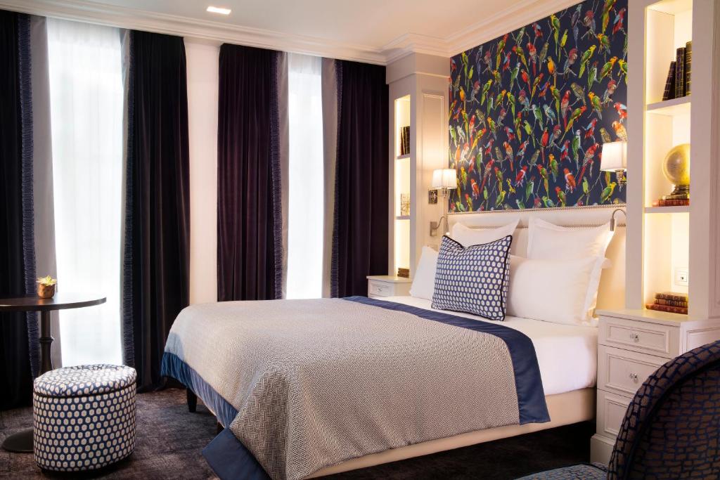 Quarto do Hotel Monge com cama de casal, papel de parede com pássaros, cortinas escuras e prateleiras embutidas com livros.
