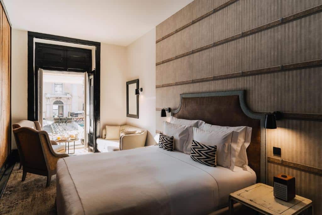 Quarto do AlmaLusa Baixa/Chiado, com cama de casal e almofadas de vários tamanhos, uma varanda à esquerda com mesa e cadeiras, e ainda duas poltronas e um pequeno sofá de dois lugares junto à cama