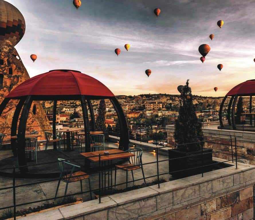 Pátio de cobertura do Carus Cappadocia com toldos vermelhos e vista sobre a cidade ao pôr do sol, com balões de ar quente a flutuar no céu.
