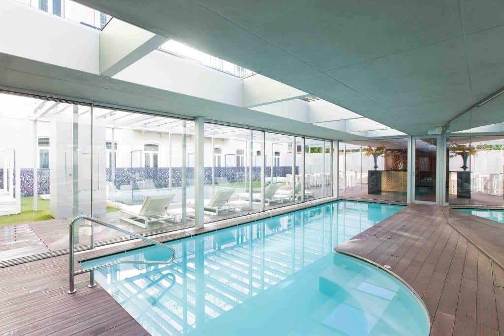 Piscina interior com deck de madeira, espreguiçadeiras e grandes janelas que deixam entrar a luz natural de um pátio exterior, no BessaHotel Liberdade.