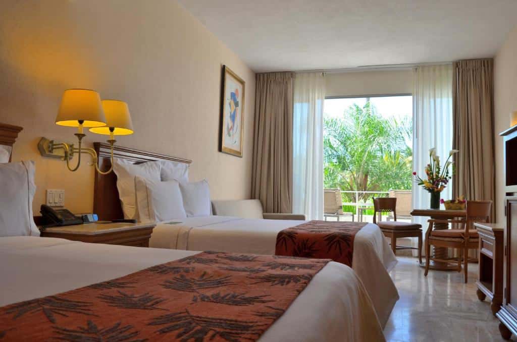 Quarto do Fiesta Americana Puerto Vallarta com duas camas, uma pequena mesa e cadeiras, candeeiros de parede e uma varanda com vista para a vegetação.