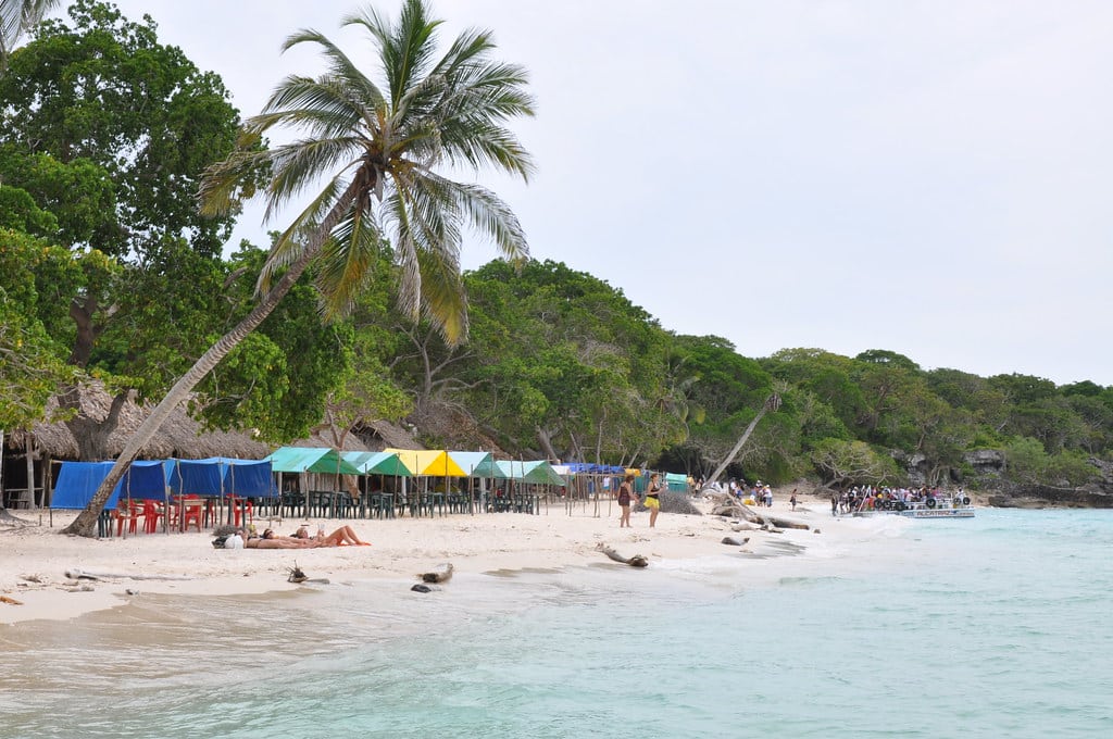 Praia tropical Playa Blanca, em Cartagena, com areia branca, água cristalina, palmeiras inclinadas e tendas coloridas junto à costa.