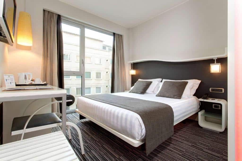 quarto do Best Western Premier Hotel Royal Santina, um dos hotéis boutique em Roma, com cama de casal grande, mesa e candeeiro de cada lado, mesa com cadeira e televisão em frente e uma janela enorme com cortinas claras e vista para a rua, com pormenores em cinza e branco