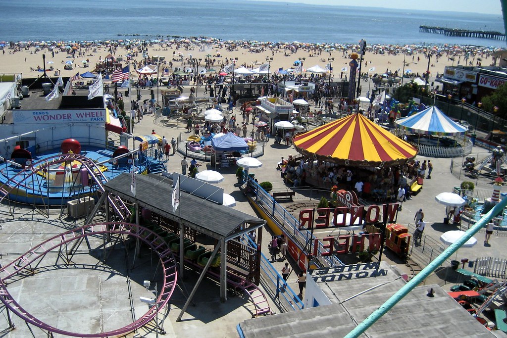 Parque de diversões movimentado em Coney Island, com roda-gigante, carrossel e uma praia cheia de gente ao sol.