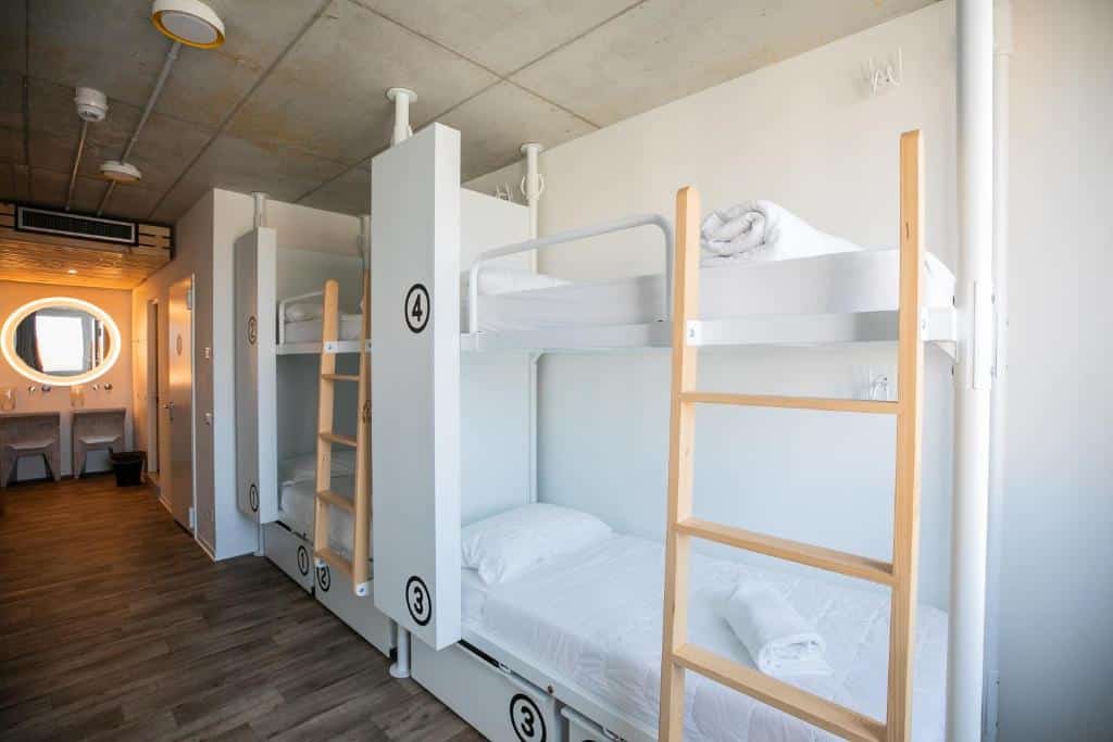 Quarto com beliches brancos e móveis de madeira clara, com artigos de higiene arrumados em prateleiras