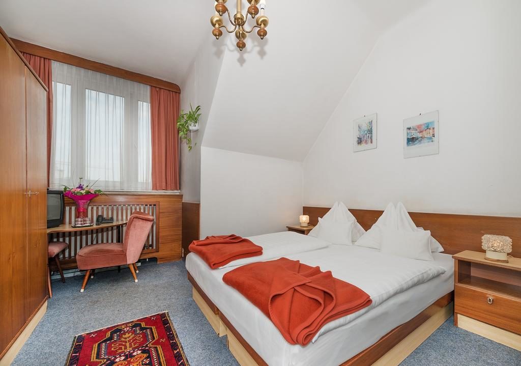 Quarto no Pension Dr. Geissler, bem arrumado, com cama de casal, cobertores cor de laranja, mobiliário de madeira, uma secretária e duas janelas com cortinados transparentes.