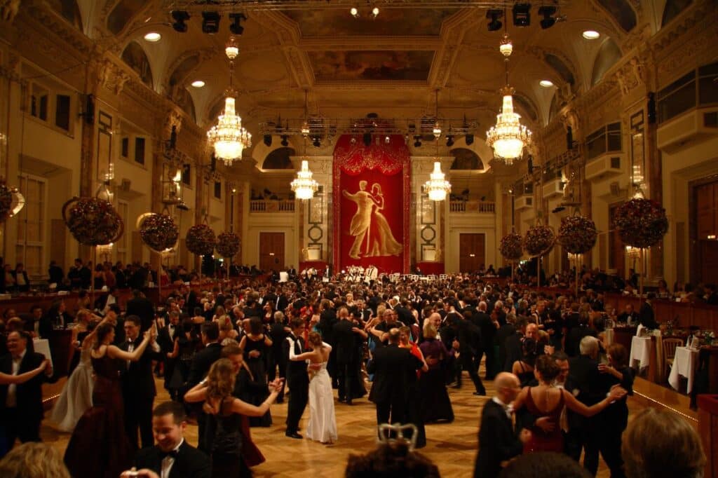 Salão do Hofburg cheio de pessoas elegantemente vestidas a dançar, sob lustres ornamentados e uma grande cortina vermelha.
