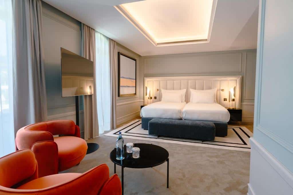 Quarto moderno do Maison Albar – Amoure, com duas camas, TV de parede, cadeiras laranja, mesa redonda e janelas amplas com cortinas.