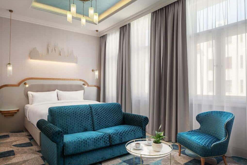 Quarto de hotel do Allure Hotel & Residence Prague, com cama de casal, sofá azul, poltrona, mesa redonda e janelas grandes.