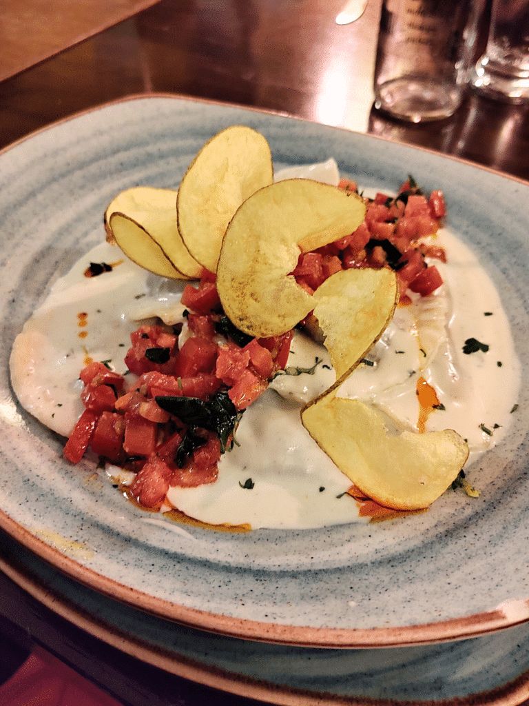 Prato quadrado azul com massa em molho branco, tomate picado e batata assada em espiral, num restaurante de Urubamba
