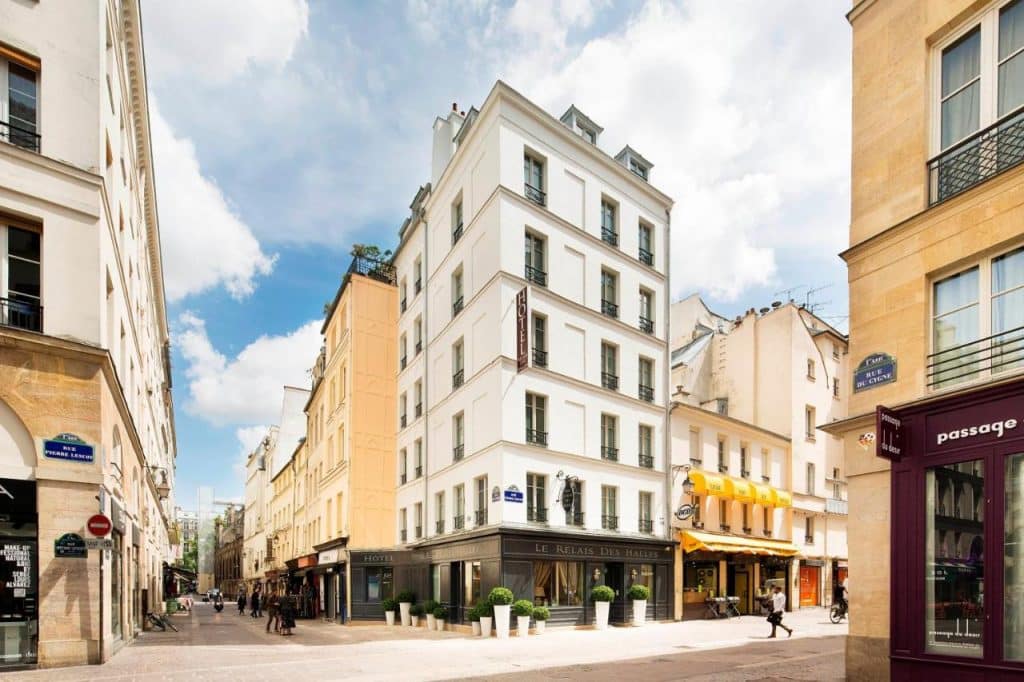 Vista de rua do Hôtel Le Relais des Halles, edifício de esquina com cinco andares, lojas e restaurantes num dia de sol.