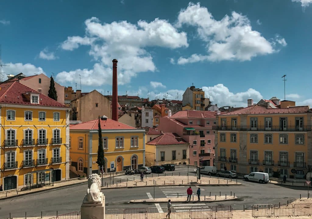 Uma praça com edifícios coloridos, carros estacionados ao longo da rua e um céu parcialmente nublado, em Lisboa.