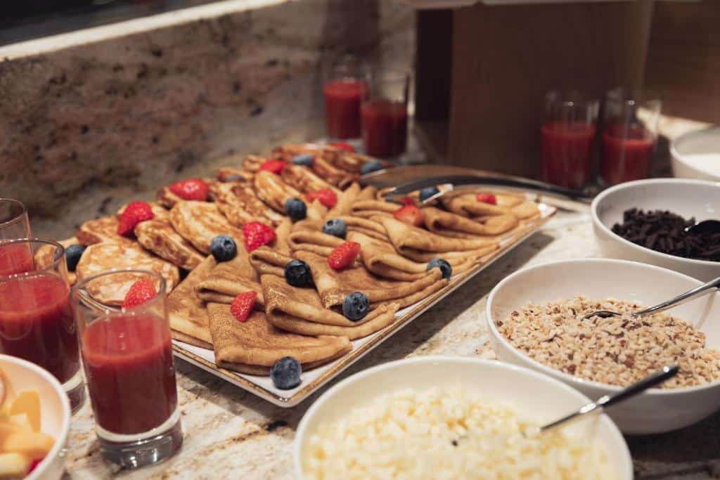 Mesa de buffet do Allure Hotel & Residence Prague com panquecas, crepes com frutos vermelhos, tigelas de cereais e copos de sumo.