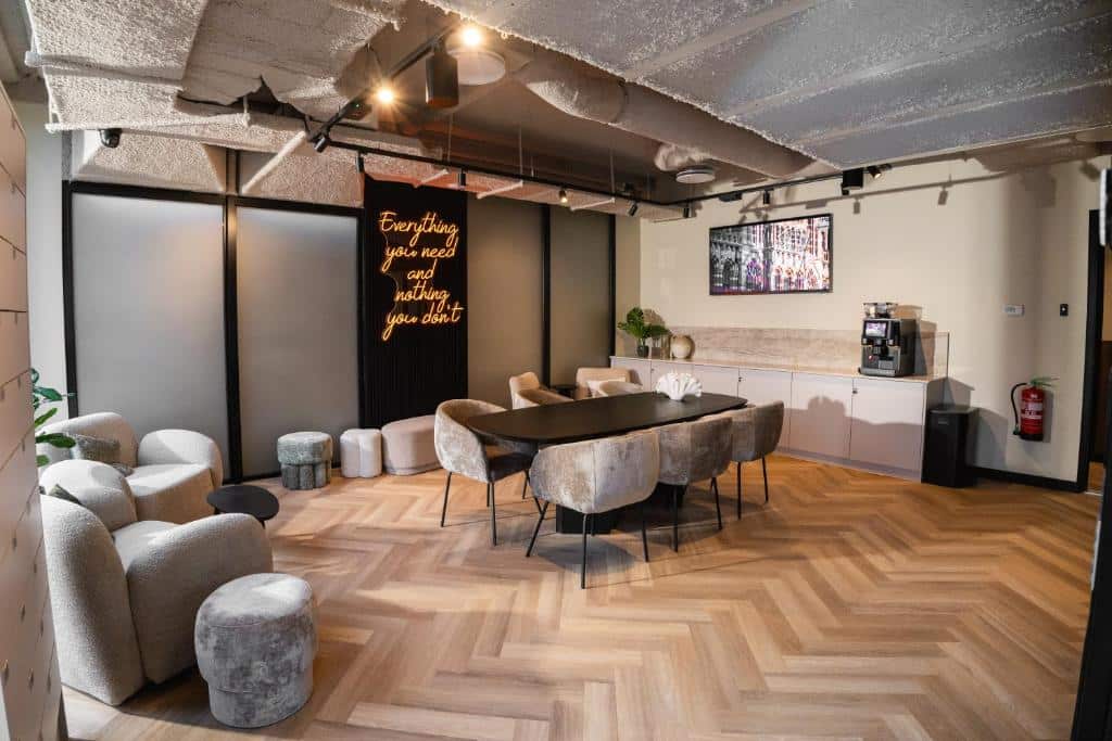Lounge moderno do Cabins Hotel Amsterdam, com uma mesa preta, cadeiras cinzentas, poltronas, pavimento de madeira e um letreiro de néon na parede do fundo.