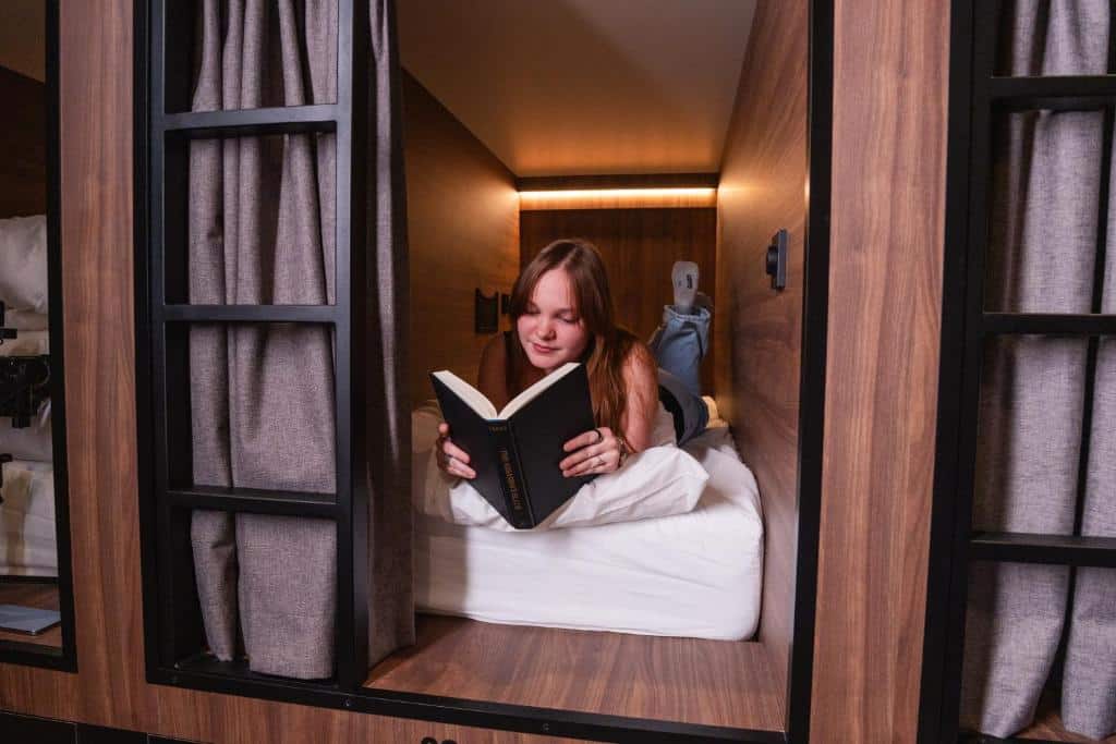 Uma pessoa deitada a ler um livro num quarto-cápsula de madeira do Cabins Hotel Amsterdam, com cortinas cinzentas parcialmente fechadas.