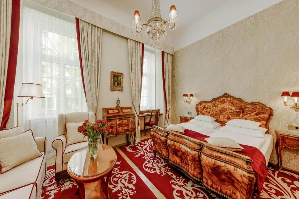 Quarto no Hotel Stefanie, com mobiliário de madeira ornamentado, apontamentos em vermelho, cama de casal, sofá, secretária e janelas grandes com cortinados.