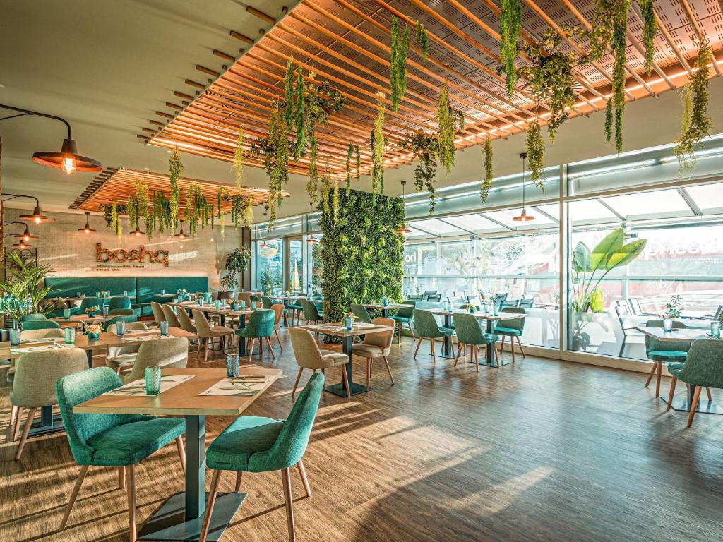 Interior do restaurante moderno do Mercure Lisboa Almada com cadeiras verdes, mesas de madeira e folhagem suspensa.