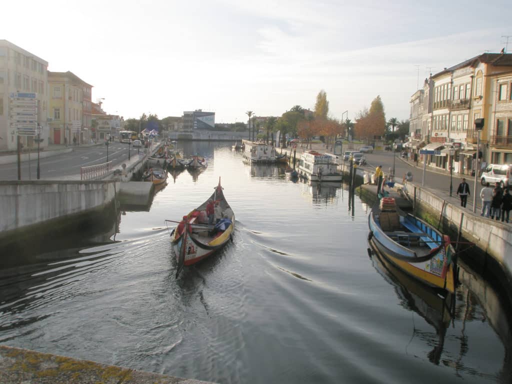 Barcos moliceiros navegam por um canal estreito em Aveiro, com edifícios e árvores de ambos os lados.