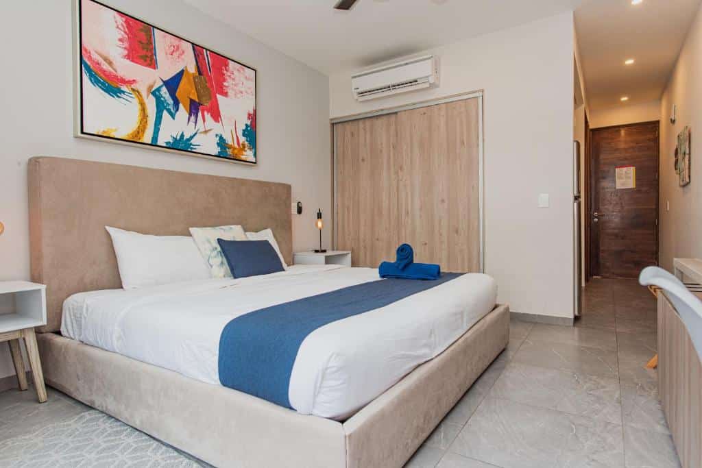 Quarto do The Gallery Condos Playa del Carmen com cama de casal, roupa de cama branca e azul, ar condicionado e quadro na parede.