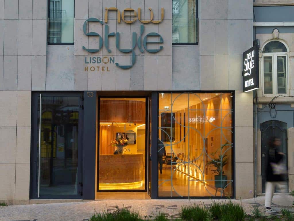 Entrada do New Style Lisbon Hotel com uma porta de vidro com desenhos geométricos e uma receção iluminada por luz quente; o edifício é cinzento.